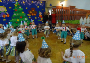 Na tle dekoracji światecznej stoją w kole dzieci z czapeczkami urodzinowymi na głowach.